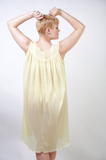 mooie jonge vrouw met kort haar en mollig lichaam het dragen van transparante Nachthemd en poseren op witte studio achtergrond alleen. mooie blonde plus size meisje in gele geplooide lingerie jurk staande. - Foto, afbeelding