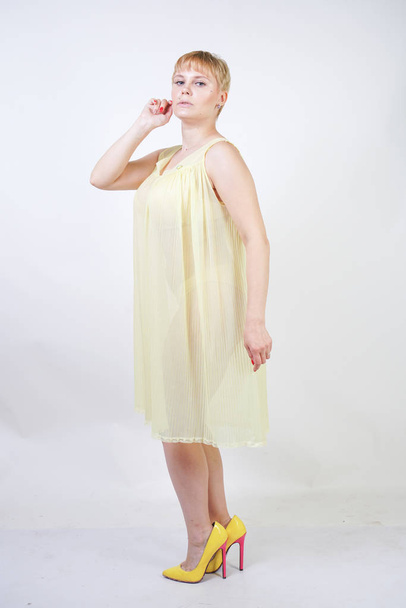 pretty young woman with short hair and chubby body wearing transparent nightgown and posing on white studio background alone. beautiful blonde plus size girl in yellow pleated lingerie dress standing. - Fotografie, Obrázek