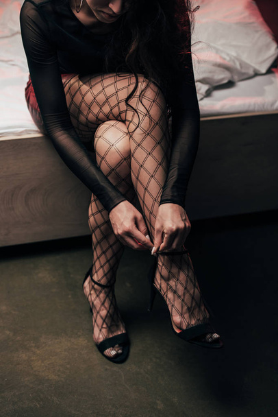 oříznutý pohled na prostitutku v mřížkových punčochách dotek obuvi při sezení na posteli  - Fotografie, Obrázek