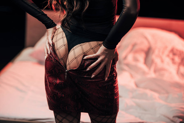 cropped view of prostitute undressing and touching buttocks in bedroom with red lights on black  - Fotografie, Obrázek