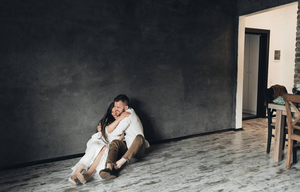 Pareja joven sentada en el suelo en casa. Hermosa pareja en vestido blanco cerca de la pared negra
.  - Foto, imagen