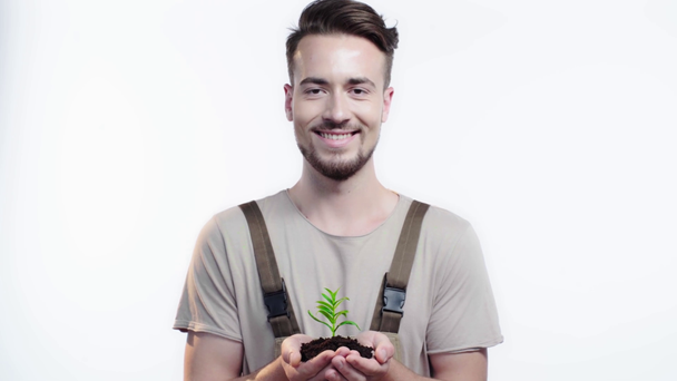 Vorderansicht des hübschen Gärtners, der eine Handvoll Erde mit grünem Spross in der Hand hält, die Augen schließt und in die Kamera lächelt - Filmmaterial, Video