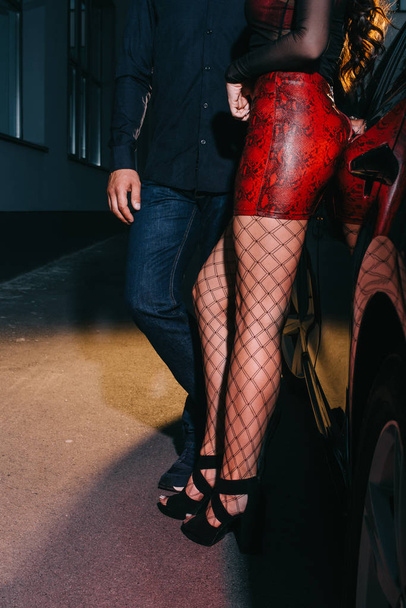 vista recortada de prostituta en falda roja y medias de malla de pie con el cliente cerca del coche
  - Foto, Imagen