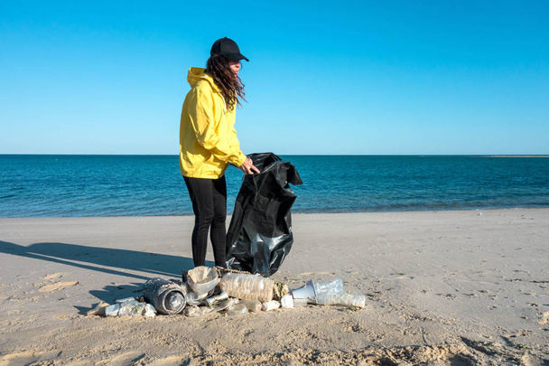 Eine Frau sammelt Müll und Plastik auf und säubert den Strand mit einem Müllsack. Freiwilliger Umweltaktivist gegen Klimawandel und die Verschmutzung der Ozeane. - Foto, Bild