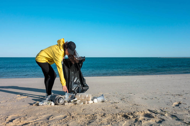 Woman picking up trash and plastics cleaning the beach with a garbage bag. Environmental volunteer activist against climate change and the pollution of the oceans. - Fotoğraf, Görsel