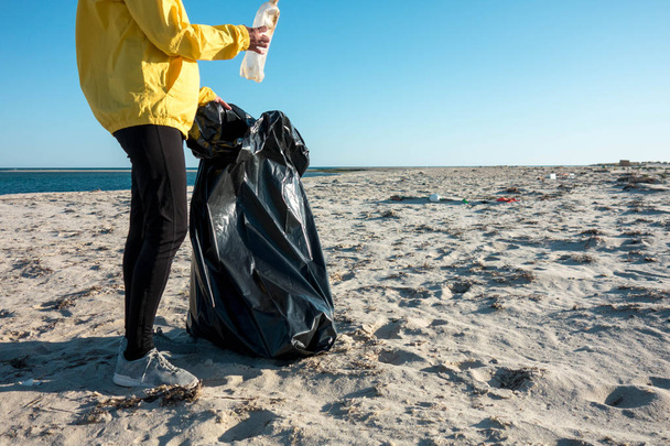 Η γυναίκα μαζεύει σκουπίδια και πλαστικά καθαρίζει την παραλία με μια σακούλα σκουπιδιών. Περιβαλλοντολογικός εθελοντής κατά της κλιματικής αλλαγής και της ρύπανσης των ωκεανών. - Φωτογραφία, εικόνα