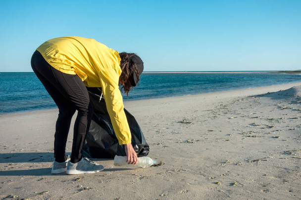 Woman picking up trash and plastics cleaning the beach with a garbage bag. Environmental volunteer activist against climate change and the pollution of the oceans. - Foto, Imagem
