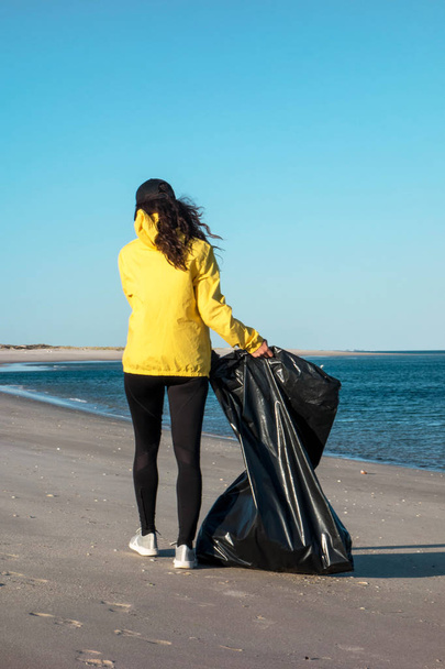 Η γυναίκα μαζεύει σκουπίδια και πλαστικά καθαρίζει την παραλία με μια σακούλα σκουπιδιών. Περιβαλλοντολογικός εθελοντής κατά της κλιματικής αλλαγής και της ρύπανσης των ωκεανών. - Φωτογραφία, εικόνα