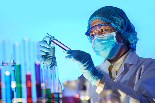 Επαγγελματίας χημικός στη μάσκα προσώπου και τα γυαλιά που αναζητούν σε δοκιμή-σωλήνα με μοβ υγρό - Φωτογραφία, εικόνα