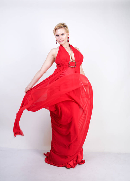 hot blonde caucasian woman wearing long red evening dress and posing on white studio background alone. fashionable adult girl with plus size body standing in stylish party clothes. - Zdjęcie, obraz