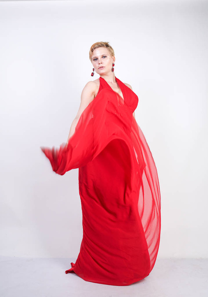 hot blonde caucasian woman wearing long red evening dress and posing on white studio background alone. fashionable adult girl with plus size body standing in stylish party clothes. - Foto, afbeelding