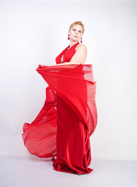hot blonde caucasian woman wearing long red evening dress and posing on white studio background alone. fashionable adult girl with plus size body standing in stylish party clothes. - Фото, зображення