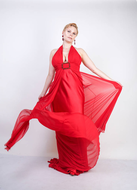 hot blonde caucasian woman wearing long red evening dress and posing on white studio background alone. fashionable adult girl with plus size body standing in stylish party clothes. - Fotó, kép