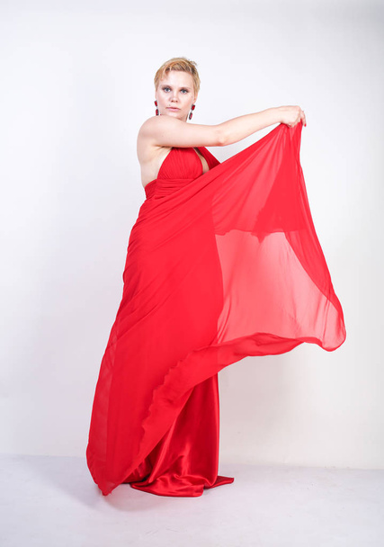 hot blonde caucasian woman wearing long red evening dress and posing on white studio background alone. fashionable adult girl with plus size body standing in stylish party clothes. - Fotoğraf, Görsel