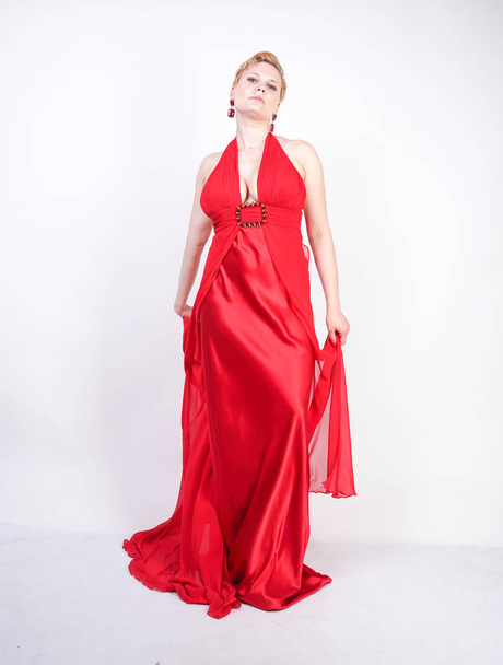 hot blonde caucasian woman wearing long red evening dress and posing on white studio background alone. fashionable adult girl with plus size body standing in stylish party clothes. - Foto, Imagem