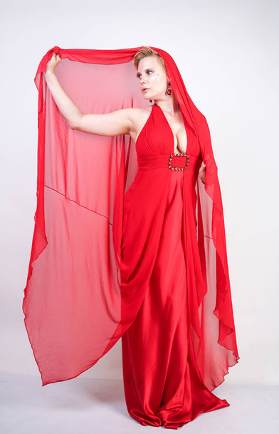 meleg szőke kaukázusi nő visel, hosszú, piros estélyi ruha, és pózol a fehér stúdió háttere egyedül. divatos felnőtt lány molett test áll stílusos party ruhák. - Fotó, kép