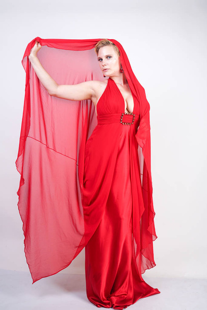 femme caucasienne blonde chaude portant longue robe de soirée rouge et posant sur fond blanc studio seul. fille adulte à la mode avec un corps de taille plus debout dans des vêtements de fête élégants
. - Photo, image