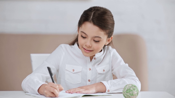 hymyilevä söpö esiteini koulutyttö tekee kotitehtäviä ja kirjoittaa kopiokirjan pöydällä
 - Materiaali, video