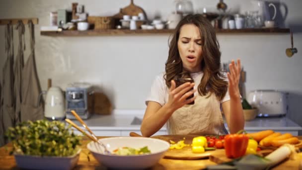 Young pretty lady receiving bad news on phone while cooking in the kitchen - Materiał filmowy, wideo