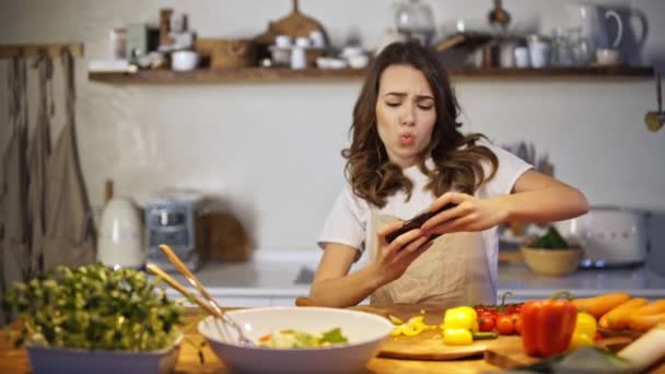 Young woman getting fun playing game on smartphone while cooking in the kitchen - Filmagem, Vídeo