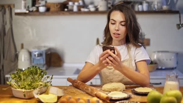Smiling young lady in apron chatting on smartphone while cooking at the kitchen - Záběry, video