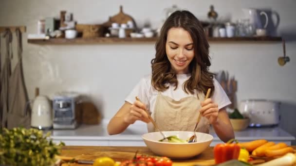 Cheerful young lady in apron making salad in the kitchen - Séquence, vidéo