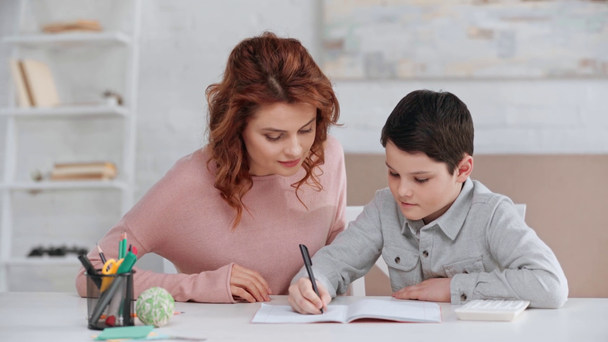 机に座り、息子が教科書を書いている間、息子の宿題を手伝う母親 - 映像、動画