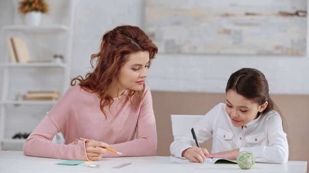 Äiti istuu pöydän ääressä ja auttaa tytärtä läksyjen kanssa, kun hän kirjoittaa oppikirjassa
 - Materiaali, video