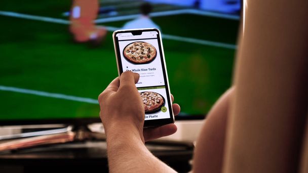 Against the background of the TV at home (at the bar), the guy (woman) orders different snacks, pizza and sweets scrolling through internet stores using mobile phones. Concept: Football, Sport,Order. - 写真・画像