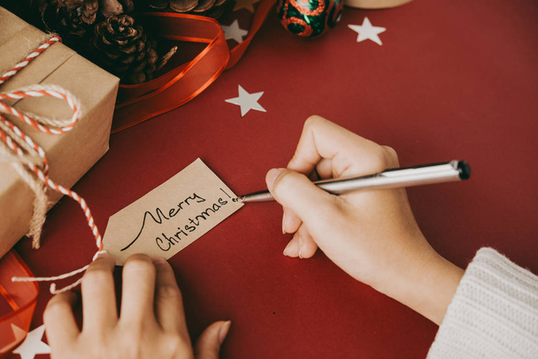 Close-up de mulher irreconhecível sentada à mesa com pano vermelho e escrevendo desejo de Natal na etiqueta enquanto prepara presentes de Natal
 - Foto, Imagem