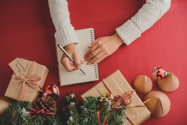 クリスマス前にメモ帳でギフトリストを構成する白いニットセーターで認識できない女性の直接のビュー、彼女はクリスマスプレゼントとクリスマスツリーとテーブルに座って - 写真・画像