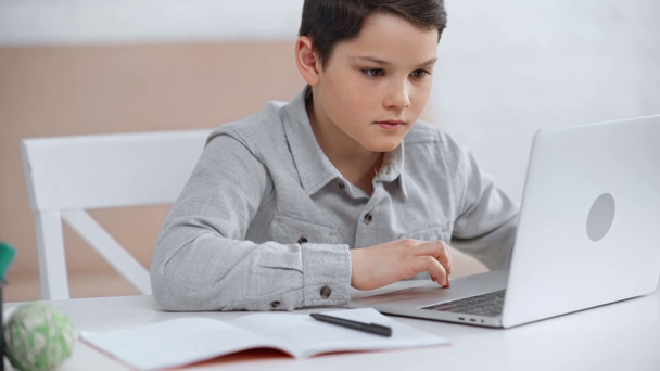 εστιασμένη Προέφηβος μαθητής κοιτάζοντας την οθόνη του φορητού υπολογιστή, γράφοντας στο βιβλίο αντιγραφής και πληκτρολογώντας στο πληκτρολόγιο - Πλάνα, βίντεο