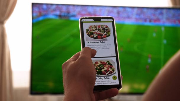 Against the background of the TV at home (at the bar), the guy (woman) orders different snacks, pizza and sweets scrolling through internet stores using mobile phones. Concept: Football, Sport,Order. - Foto, Imagem