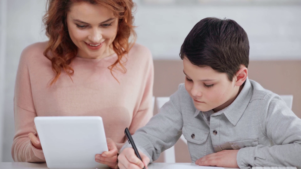 smiling mom showing digital tablet screen to son while he writing in copy book - Footage, Video