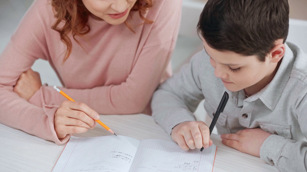 mother holding pencil and helping son with homework while he writing on copy book at desk - Footage, Video
