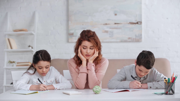 tired mother sitting at desk and looking at kids writing in copy books while doing homework - Video