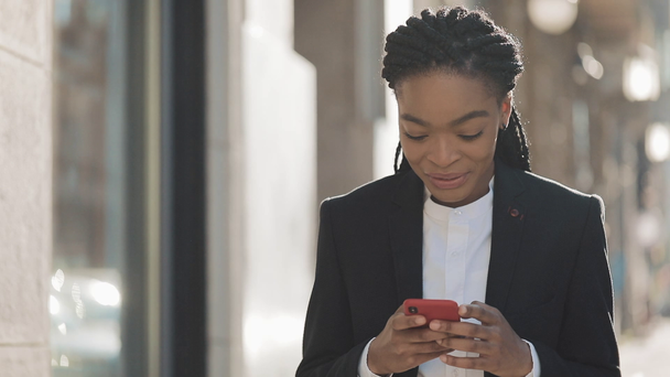Attractive businesswoman using smartphone walking on the street near business centre. Black Stylish. Dreadlocks. Female professional in suit having good news on smartphone. - Footage, Video