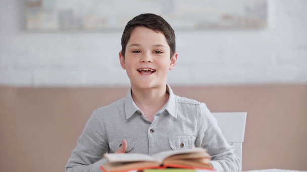 front view of smiling preteen boy sitting at desk, showing thumbs up and rock signs, making funny faces and putting book on head - Footage, Video
