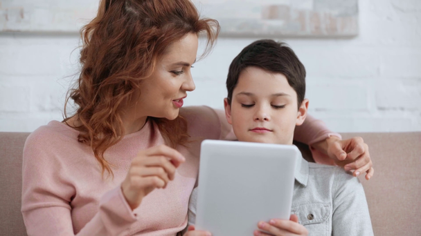 улыбающаяся мать обнимает сына и использует цифровой планшет, сидя на диване в гостиной
 - Кадры, видео