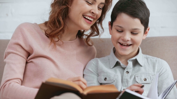uśmiechnięta matka trzyma książkę i pomaga synowi z pracy domowej siedząc na kanapie - Materiał filmowy, wideo