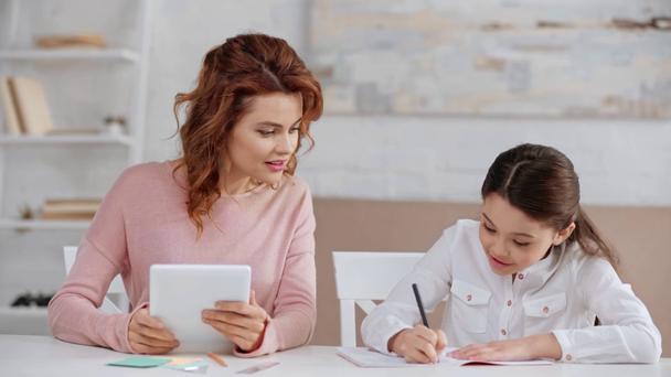 schöne lächelnde Mutter nutzt digitales Tablet am Schreibtisch, während Tochter Hausaufgaben macht - Filmmaterial, Video
