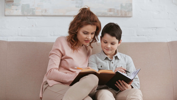 sonriente madre sentada en un sofá y leyendo libro mientras su hijo escribe en un libro de texto y sonríe en la sala de estar
 - Metraje, vídeo