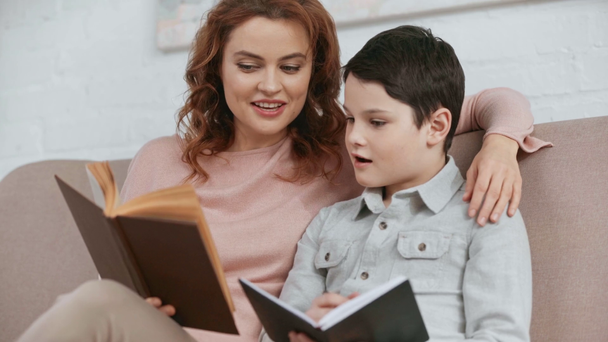 Lächelnde Mutter sitzt auf dem Sofa, umarmt Sohn und liest Buch, während er im Lehrbuch schreibt - Filmmaterial, Video