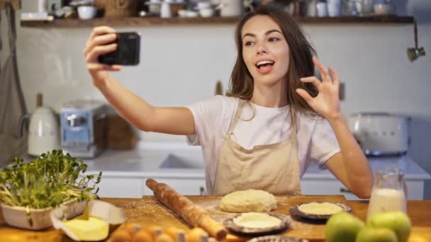 Attractive young woman in apron taking selfie photo on smartphone while cooking at the kitchen - Felvétel, videó