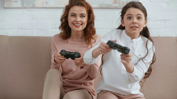 KYIV, UKRAINE -  APRIL 15, 2019: front view of mother and daughter holding joysticks and laughing while playing video game on sofa in living room - Video