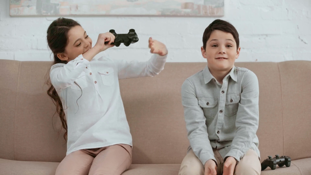 KYIV, UKRAINE -  APRIL 15, 2019: two excited kids holding joystick and playing video game while sitting on sofa in living room - Πλάνα, βίντεο