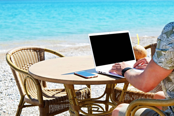 Young man working on his laptop at the beach at tropical destination. Office on the beach concept. - Photo, Image