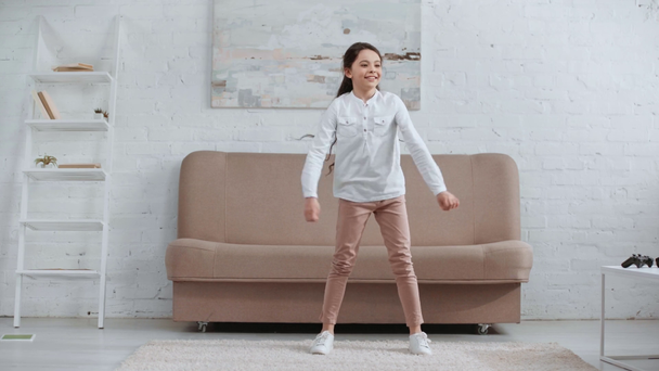 KYIV, UCRANIA - 15 DE ABRIL DE 2019: vista completa del feliz niño sonriente bailando en la alfombra cerca del sofá en la sala de estar
 - Metraje, vídeo