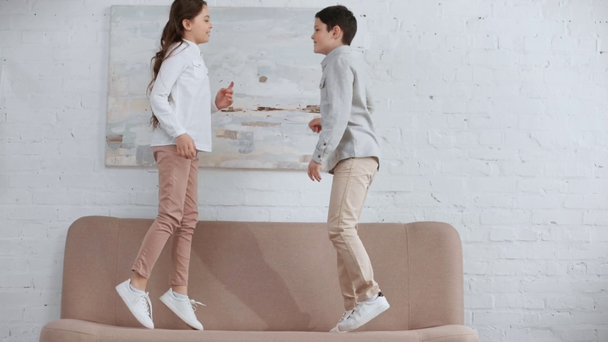 full length view of two happy smiling preteen kids jumping and dancing on sofa in living room - Footage, Video