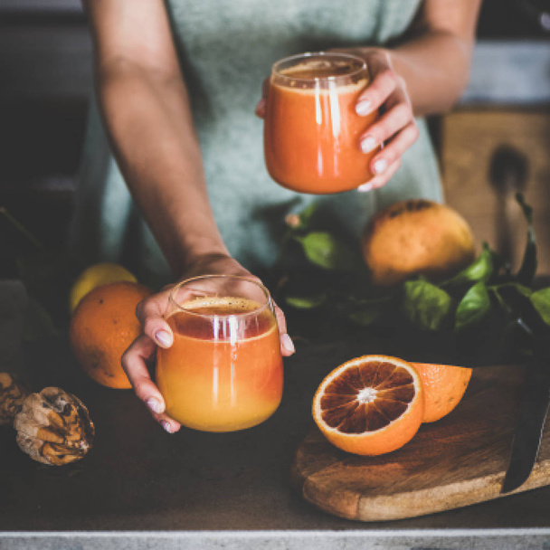 Young female holding two glasses of freshly squeezed blood orange juice or smoothie in hands near concrete kitchen counter, square crop. Healthy lifestyle, vegan, vegetarian, alkaline diet concept - Φωτογραφία, εικόνα